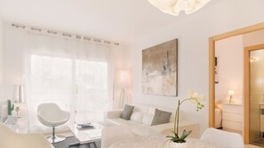 Une chambre d’hôtel moderne avec un canapé blanc, une table ronde et une grande fenêtre ornée de rideaux légers.