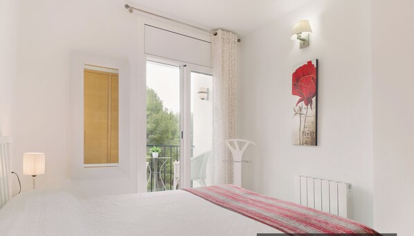 A bedroom with a bed, a window, a door, a radiator, and a wall-mounted light fixture.