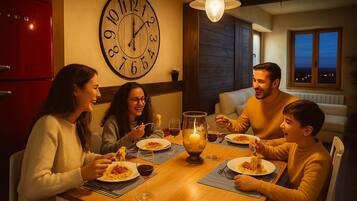 Una familia cenando en una mesa de madera con un reloj de pared grande, una vela encendida y vistas del cielo nocturno a través de una ventana.