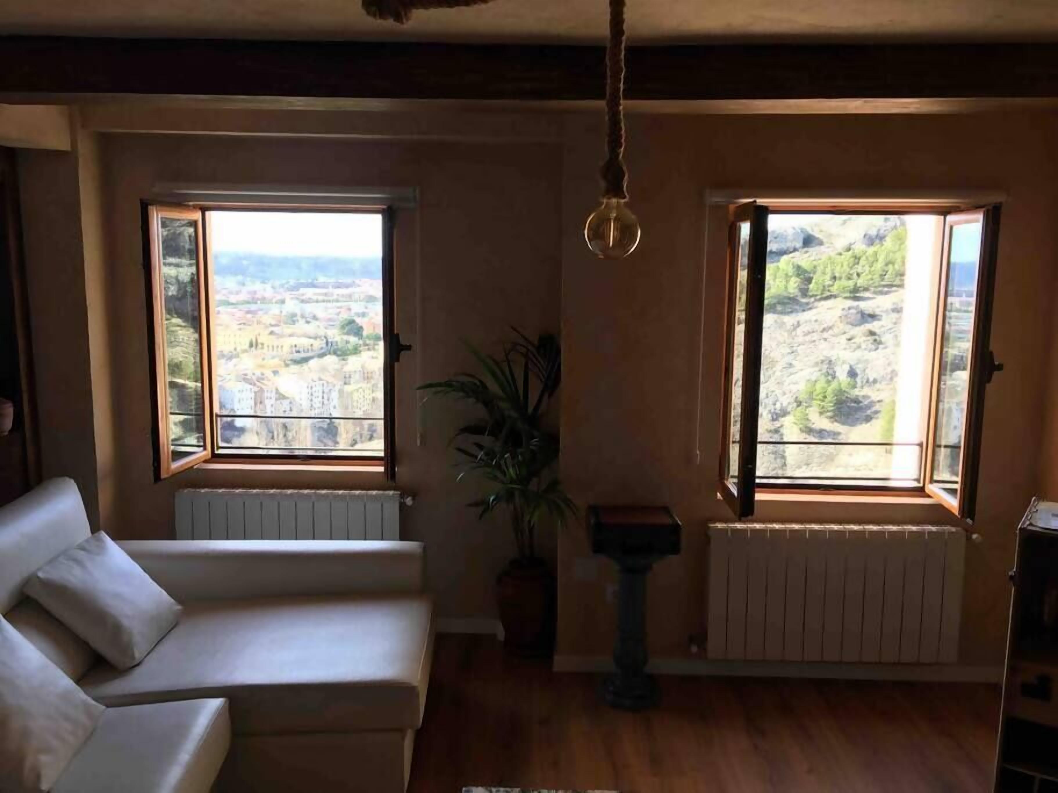 Salón con sofá blanco, dos ventanas con vistas a la ciudad, lámpara colgante y radiador.