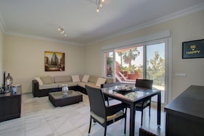 A modern living room with a sectional sofa, a glass dining table, wicker chairs, a coffee table, and a painting on the wall.