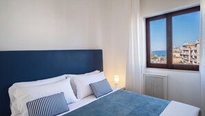 A bed with a dark headboard, white bedding, and striped pillows. A window with a view of buildings and a blue sky is visible. - Nice and sunny apartment located on the main street of Sorrento (Sorrento)
