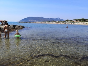 A beach with clear shallow waters, rocky shore, and a mountain in the background.