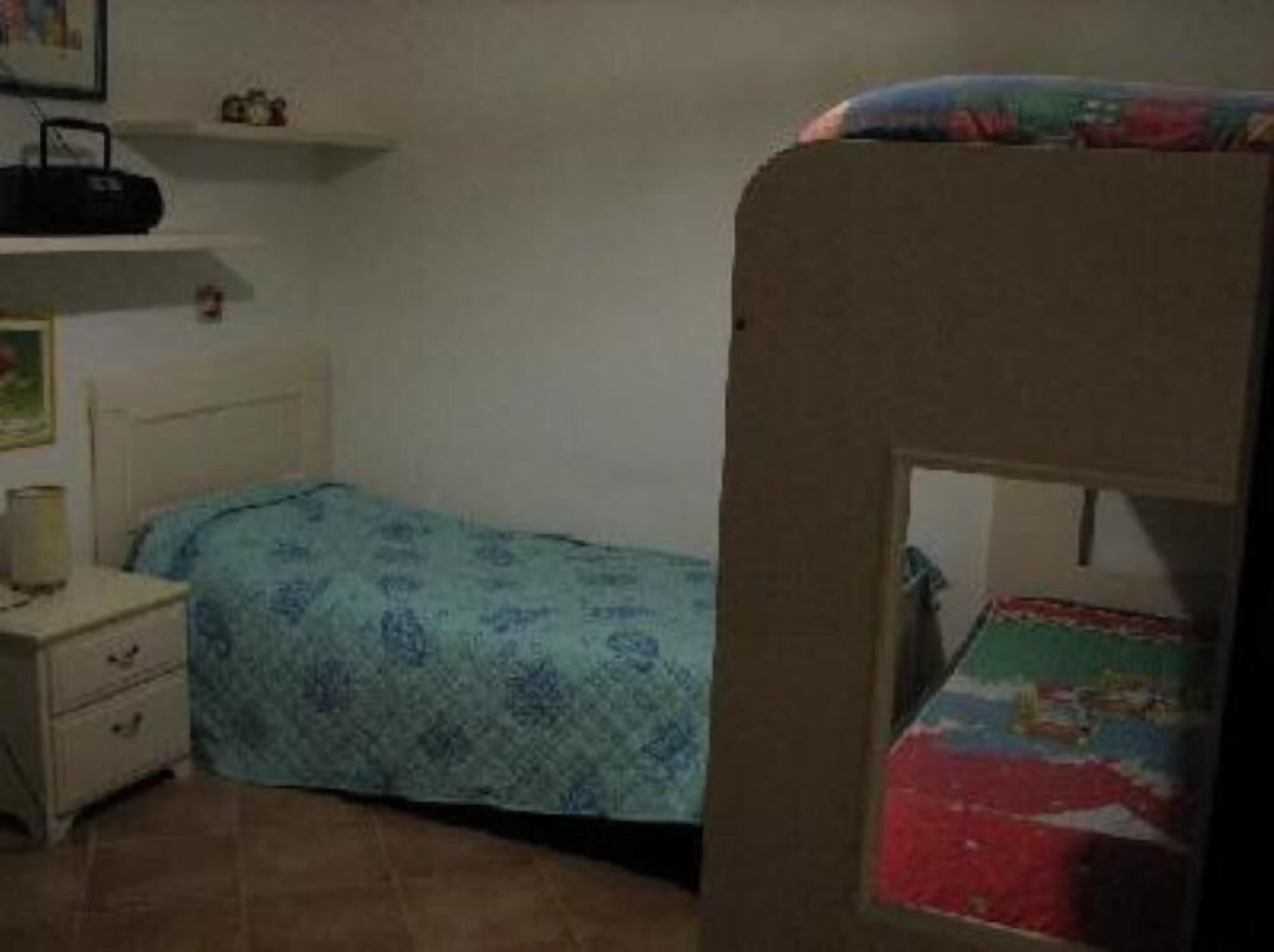 Una habitación pequeña y poco amueblada con una cama individual, una mesita de noche, un estante con una radio y un cuadro colgado en la pared.