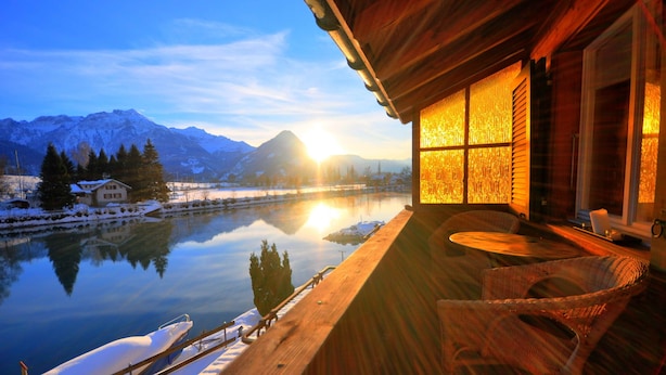 Eine Holzjagdhaus mit Balkon mit Blick auf einen ruhigen See und verschneite Berge im Hintergrund.
