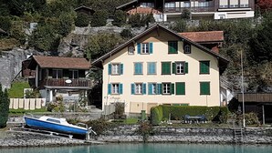 Ein zweistöckiges Gebäude mit Balkon, ein Boot liegt am Ufer und eine Steinmauer im Vordergrund.