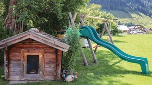 Una cabaña de madera con una puerta metálica, un tobogán verde y un columpio en un área cubierta de césped, con árboles y una montaña al fondo.
