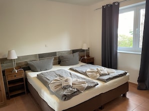 A double bed with grey bedding, two bedside tables with lamps, a window with curtains, and a view of greenery outside. - Suite Friedhelm 9a UR with terrace (SO) - Usedom Suites 09***, beach 100 m (Karlshagen)