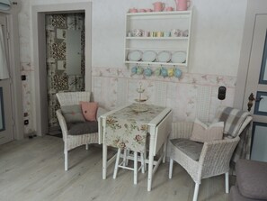 A dining area with wicker chairs, a table with floral tablecloth, and a shelf with kitchenware. - Apartment country house style - Haus Bindseil (Altenau)
