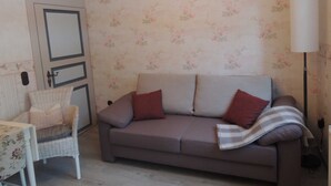 Una habitación con un sofá gris, una silla de mimbre, una lámpara blanca y papel tapiz floral.