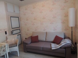 A room with a grey sofa, a wicker chair, a white lamp, and floral wallpaper. - Apartment country house style - Haus Bindseil (Altenau)