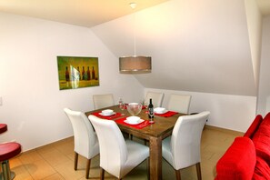 Uma sala de jantar com uma mesa de madeira posta para seis pessoas, cadeiras brancas, um lustre e um quadro na parede.