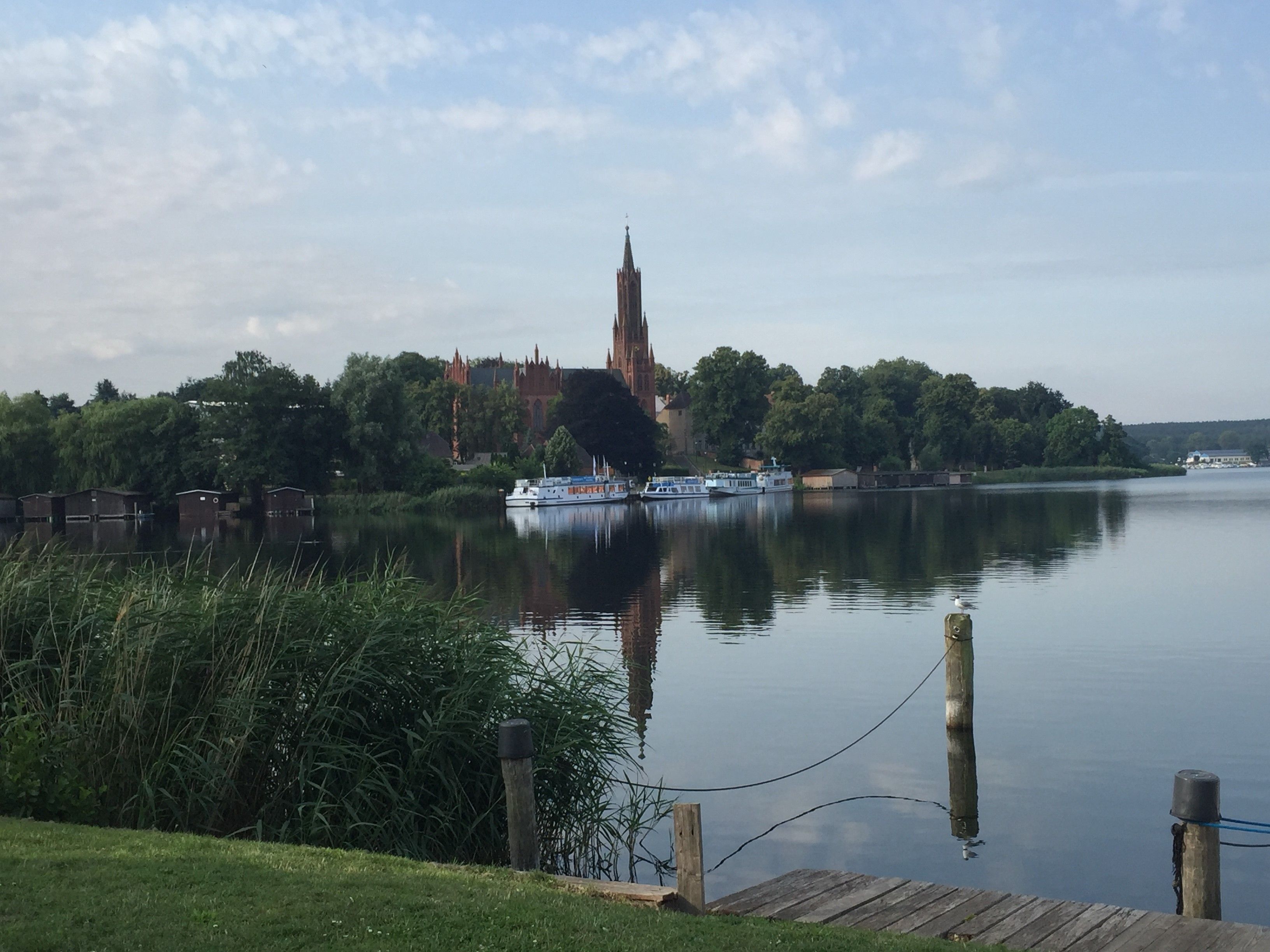 En flod med en båd, der ligger til anker på højre side, en træbro i forgrunden og en stor kirke med et højt spir i baggrunden.