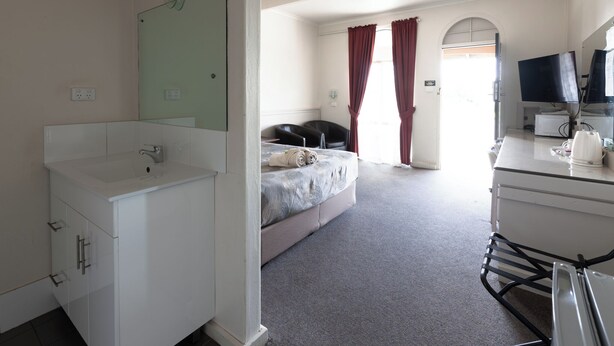 A hotel room with a white bathroom, a bed with a grey bedspread, a desk with a computer and a flat-screen TV, and a window with red curtains. - The Argent Motel (Broken Hill)