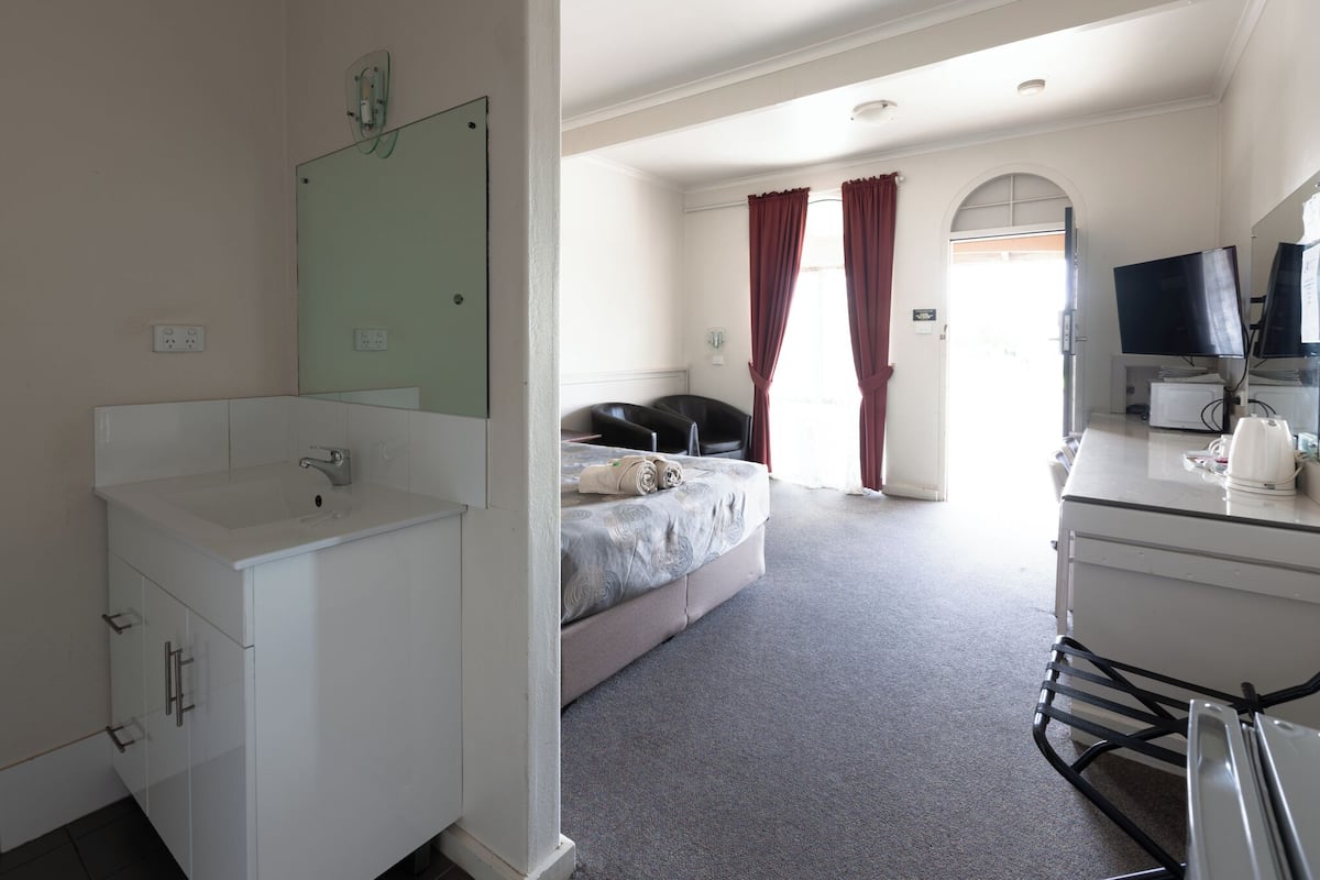 Ein Hotelzimmer mit weißem Badezimmer, einem Bett mit grauer Bettdecke, einem Schreibtisch mit Computer und Flachbildfernseher sowie einem Fenster mit roten Vorhängen.