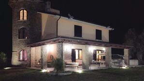 Ein zweistöckiges Steinhaus mit einem Turm, das eine Terrasse mit Sitzgelegenheiten und einen gut beleuchteten Eingangsbereich besitzt.
