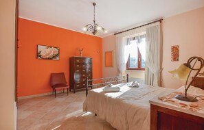Une chambre avec des murs orange, un lit avec une literie blanche, une commode en bois, une chaise et une fenêtre avec des rideaux.