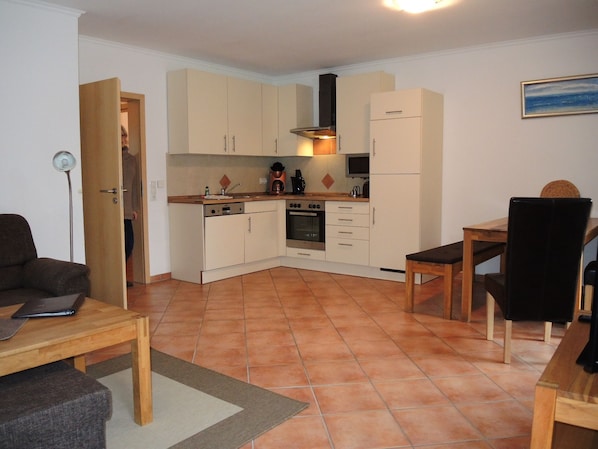 A compact kitchen with white cabinets, a stove, and a refrigerator. There is a dining table with chairs and a small rug on the floor. - Suite Max with terrace 27 u (SA) - Usedom Suites 27***, beach 100 m (Karlshagen)