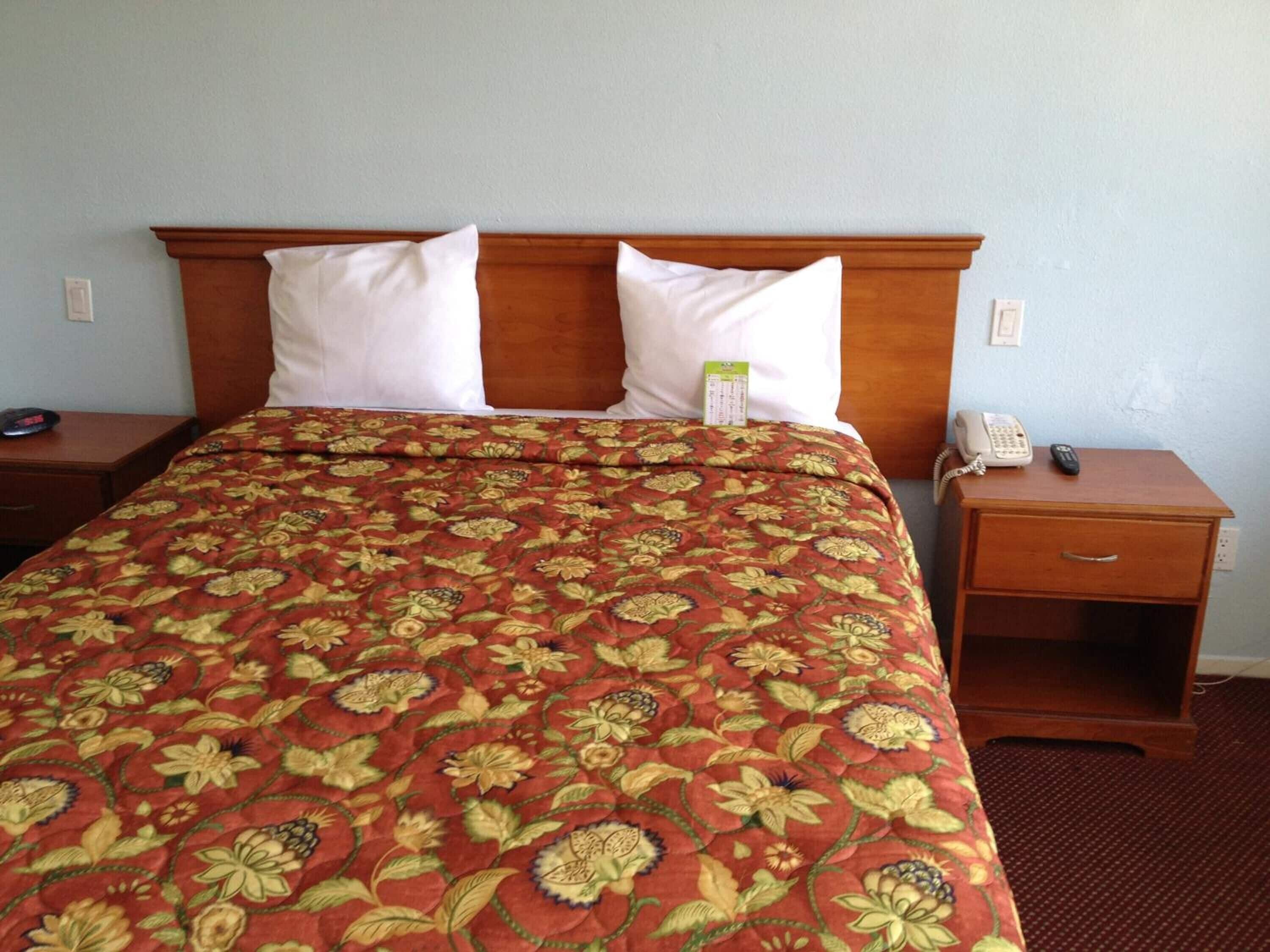 A hotel room with a large bed, two white pillows, a floral-patterned bedspread, a wooden headboard, a bedside table with a phone and remote control, and a small drawer.