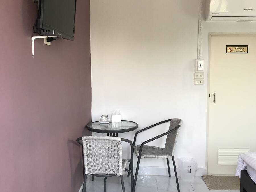 A small room with a TV mounted on the wall, a round table with two chairs, a bed, and a door with a 'No Smoking' sign.