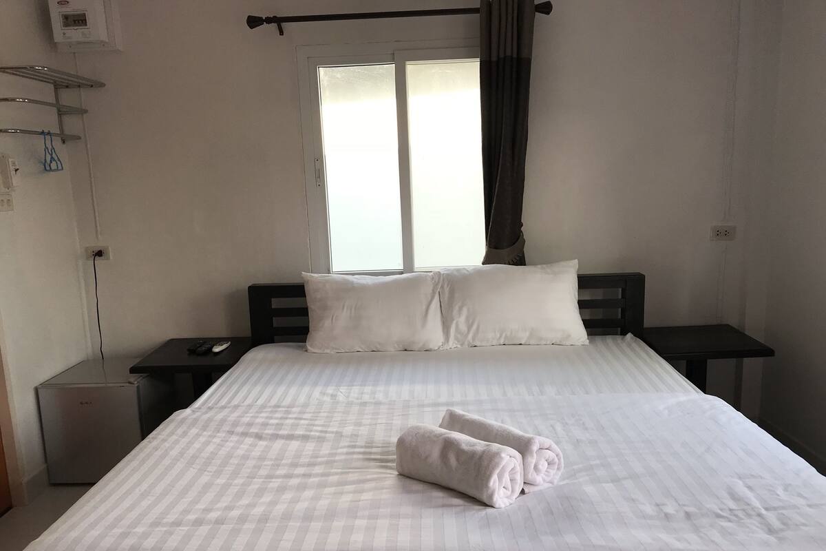 Ein ordentlich bezogenes Bett mit weißen Leinentüchern und zwei Kissen, ein Nachttisch mit Fernbedienung und ein Fenster mit Vorhängen.