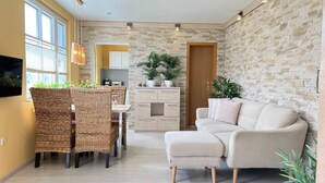 A modern living room with a sofa, dining table, wicker chairs, a kitchen area, and stone wall decor. - Apartment 3 No. - Villa Trautes Heim (Heringsdorf)