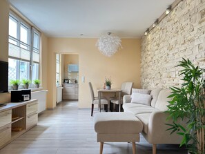 A modern living room with a sofa, ottoman, dining table, and chairs. There is a kitchen area in the background with a microwave and a TV. The room has large windows and a stone wall.
