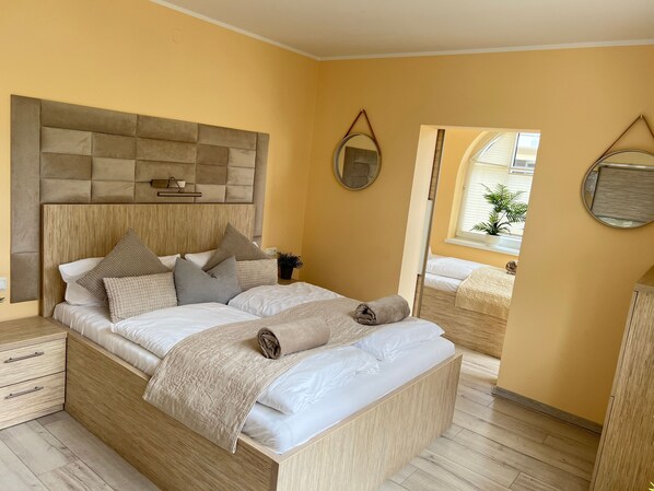 Un dormitorio con cabecera de madera, cama con sábanas blancas y varias almohadas, mesita de noche con lámpara y espejo en la pared.