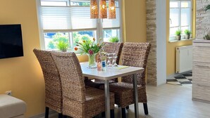 Un coin repas avec des chaises en osier, une table agrémentée d’un vase de fleurs et une fenêtre avec des stores.