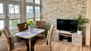 A room with a stone wall, a TV on a wooden stand, wicker chairs, a table with a vase of flowers, and large windows. - Apartment 3 No. - Villa Trautes Heim (Heringsdorf)