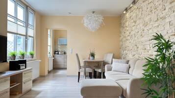 Un salon moderne comprenant un canapé, un pouf, une table à manger et des chaises. On aperçoit, au fond, un coin cuisine avec un four à micro-ondes et un téléviseur. La pièce est dotée de grandes fenêtres et d’un mur en pierre.