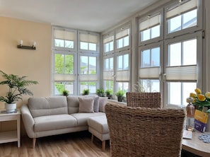 Een kamer met een bank, rieten stoelen, potplanten en een tafel met boeken en een fles.