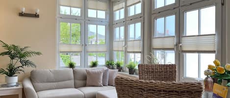 A room with a sofa, wicker chairs, potted plants, and a table with books and a bottle.