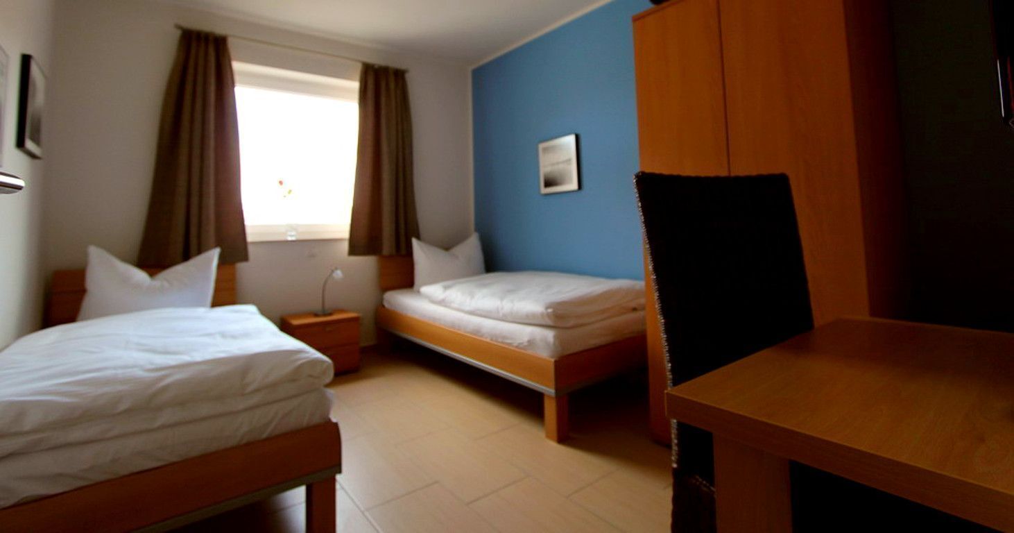 Habitación de hotel con dos camas individuales, una mesa de madera, una mesita de noche, una ventana con cortinas y una pared azul.