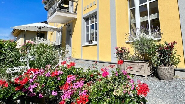 Un edifici groc amb un balcó, grans finestres i un jardí ple de flors.