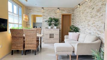 Ein modernes Wohnzimmer mit Sofa, Esstisch, Korbstühlen, einer Küchenzeile und Wandgestaltung mit Steinoptik.