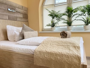 Een keurig opgemaakt bed met een beige sprei en kussens, een houten hoofdbord en een raam met plantenpotten.