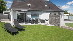 A modern house with a patio, two lounge chairs, a dining table, and a barbecue grill.