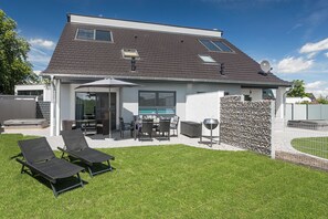 A modern house with a patio, two lounge chairs, a dining table, and a barbecue grill.