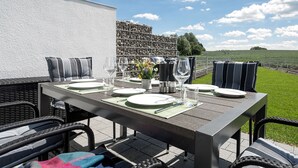 An outdoor dining area with a table set for four, surrounded by wicker chairs, a white wall, and a view of a green field.