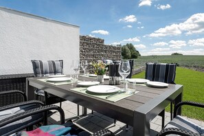 An outdoor dining area with a table set for four, surrounded by wicker chairs, a white wall, and a view of a green field.