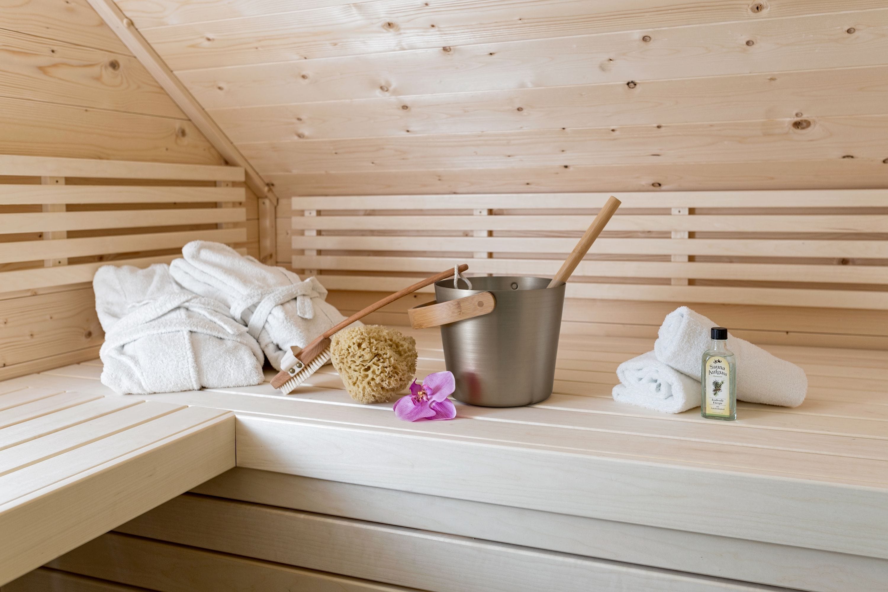 Eine Holzsauna mit Handtüchern, einer Holzbürste, einem Metallkübel, einem Schwamm und einer Lotionflasche auf einer Sitzbank.
