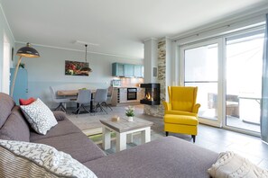 A modern living room with a sofa, armchair, and a small table. There is a kitchen area with cabinets and a fireplace.