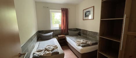 A bedroom with two single beds, a window with curtains, a bedside table, a lamp, and a wardrobe.