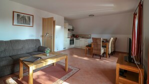 Ein Wohnzimmer mit einer Couch, einem Couchtisch und einer TV-Konsole. Im Hintergrund ist eine Küche mit einem Esstisch und Stühlen zu sehen.