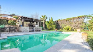 Una piscina con cubierta de piedra, zona de barbacoa con muro de piedra y pérgola con mobiliario exterior.