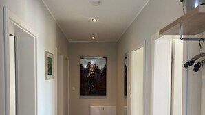 A hallway with white walls, a central painting, recessed lighting, and a potted plant on the right. - Villa Grande / Luxus-EG-Fewo ENJOY - Villa Grande / Luxus-EG-Fewo ENJOY (WE 2) (Göhren-Lebbin)