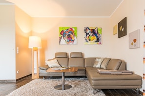 Un salon moderne avec un canapé en cuir marron, une table basse en bois et un tapis moelleux. Deux tableaux abstraits ornent le mur, et une lampe sur pied est placée dans un coin.