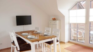 Un coin repas avec une table en bois, des chaises blanches, un téléviseur et une vue sur l’extérieur.