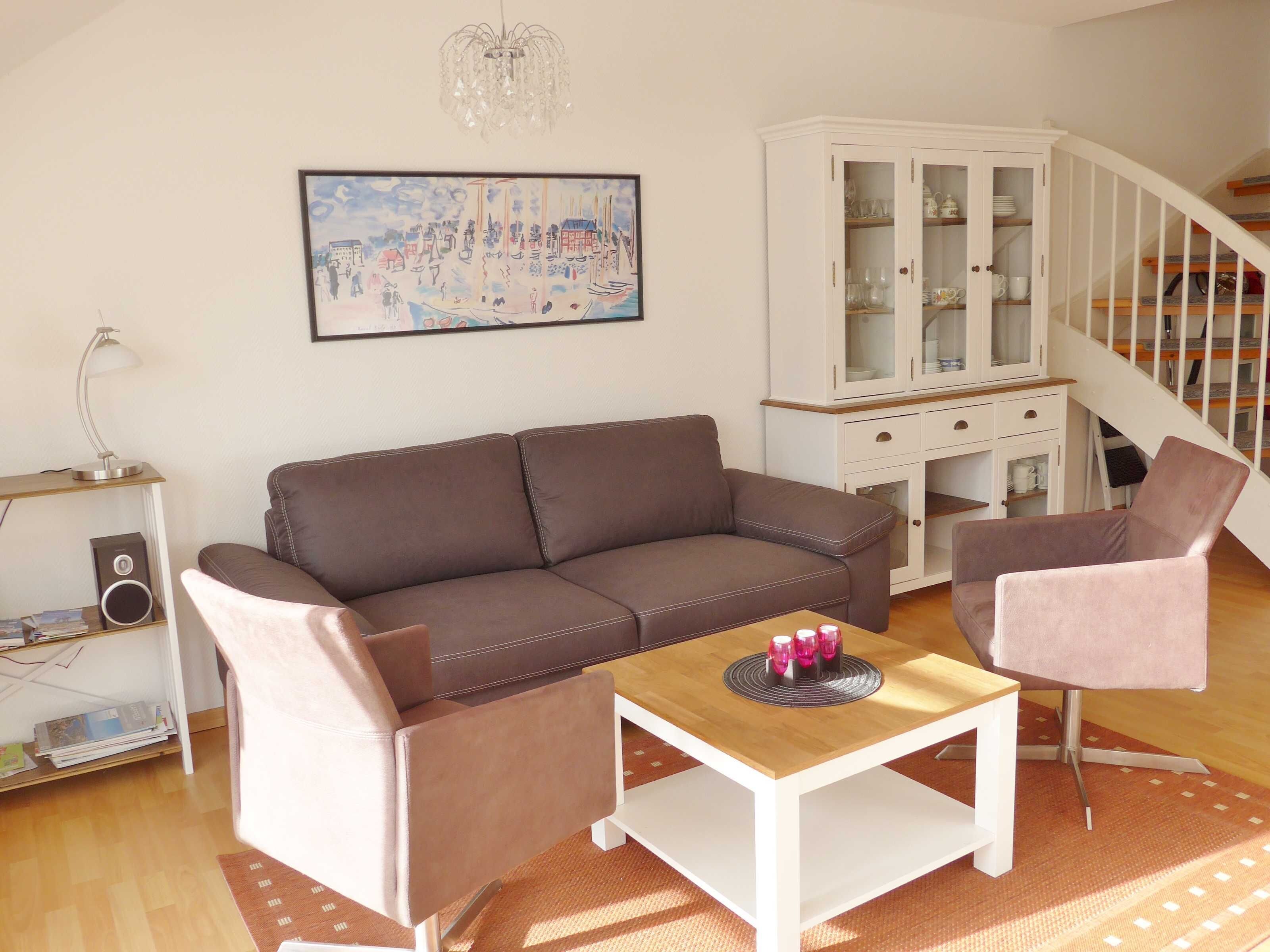 Ein Wohnzimmer mit braunem Sofa, einem rosafarbenen Sessel, einem Holztisch, einer weißen Kommode und einem Bild an der Wand.